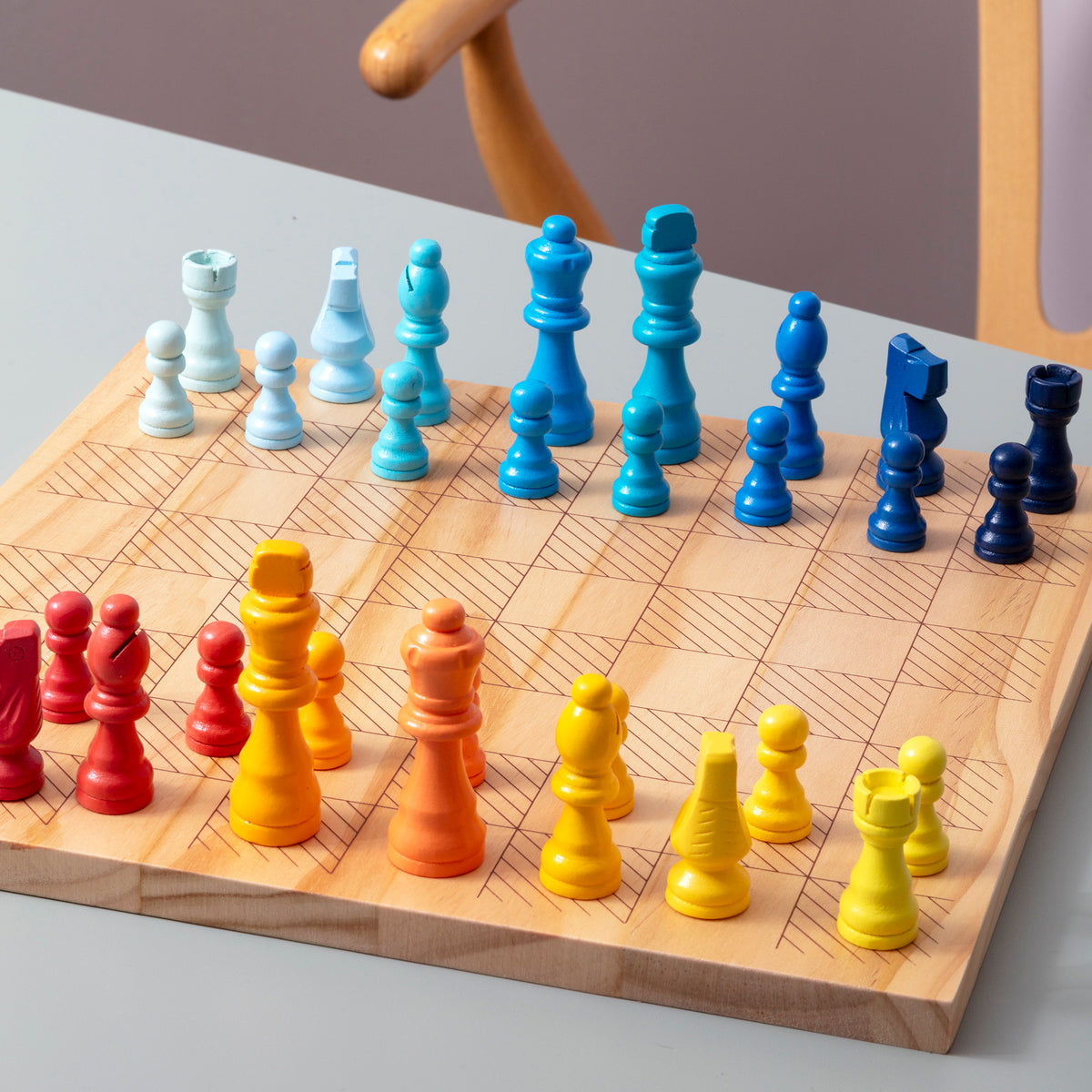 A wooden chessboard with colorful gradient chess pieces arranged in two rows, photographed on a light tabletop with part of a chair visible in the background.